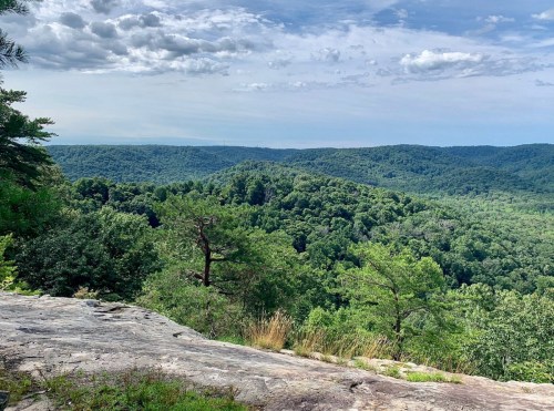A scenic view of lush green mountains and valleys under a partly cloudy sky, with rocky foreground.