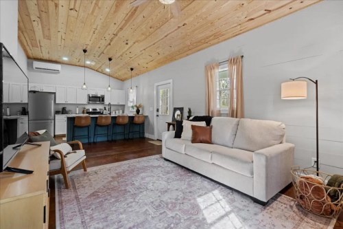 Cozy living room with a light sofa, wooden ceiling, and open kitchen area featuring bar stools and a decorative rug.