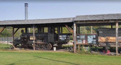 Historic lumber mill with a vintage steam engine and informational display under a wooden shelter. Green grass in foreground.