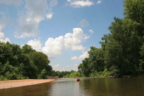 A serene river scene with lush green trees, fluffy clouds, and kayakers enjoying a sunny day.