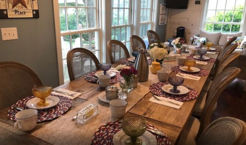A beautifully set dining table with colorful dishes, glasses, and floral centerpieces, surrounded by large windows.