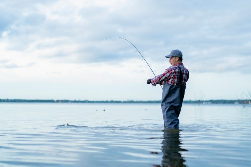 A man in waders stands in shallow water, casting a fishing line under a cloudy sky.