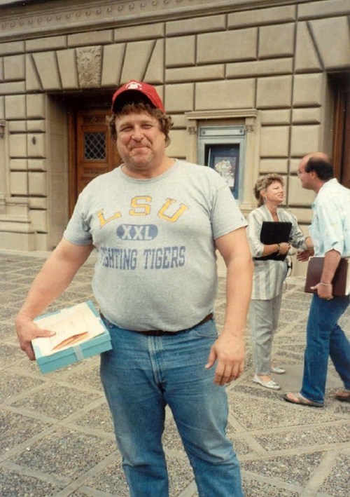 A man in a gray LSU t-shirt and red cap stands outside a building, holding a box and smiling.