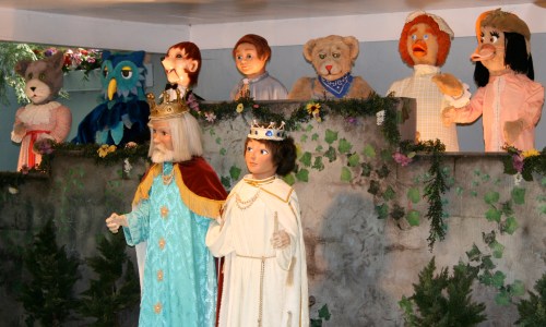 A colorful puppet show featuring various characters, including a king, queen, and animals, set against a decorated backdrop.