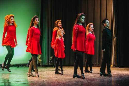 A group of dancers in red dresses perform on stage, showcasing traditional dance moves with a vibrant backdrop.