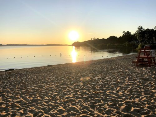 Sunset over a calm lake, reflecting golden light on the water, with a sandy beach and a lifeguard chair in the foreground.