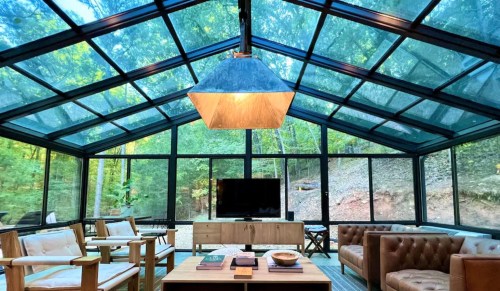 A modern glass greenhouse with a cozy seating area, wooden furniture, and a large TV, surrounded by trees.