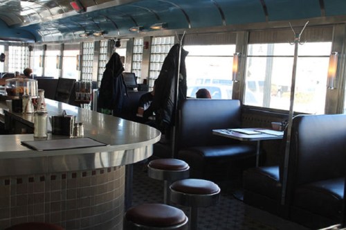 A retro diner interior featuring a curved counter, black booths, and large windows with natural light.
