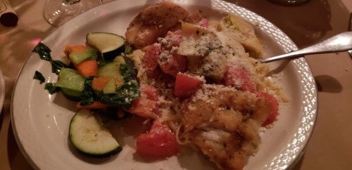 A plate of pasta topped with cheese, accompanied by sautéed vegetables and a piece of fried fish.