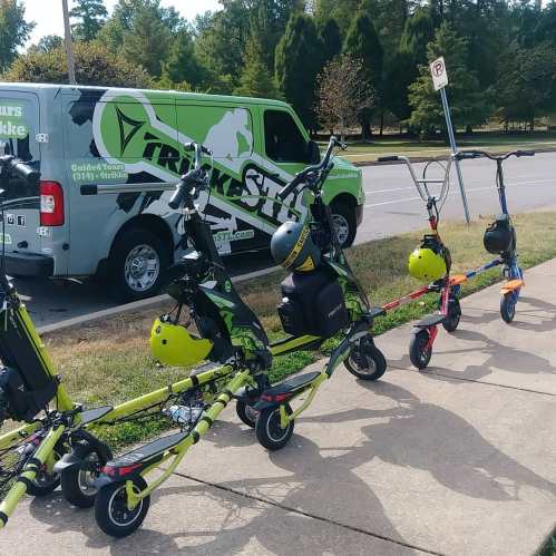 A row of colorful scooters with helmets parked beside a green van labeled "Trike STL" in a park setting.