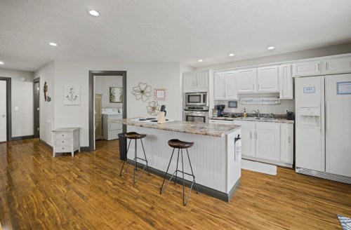 Modern kitchen with white cabinets, granite island, and wooden floors, featuring bar stools and stainless steel appliances.