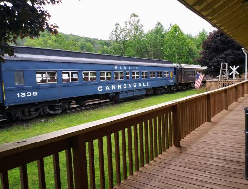 A blue train car labeled "CANNONBALL" parked beside a wooden deck, surrounded by greenery and an American flag.