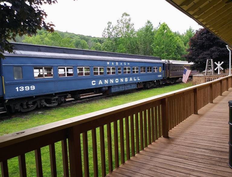 A blue train car labeled "CANNONBALL" parked beside a wooden deck, surrounded by greenery and an American flag.