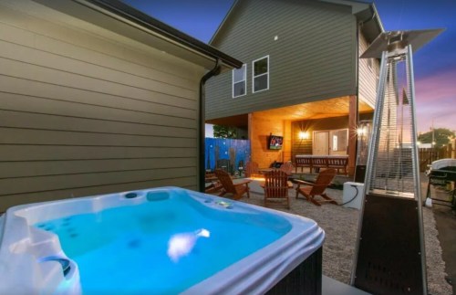 A hot tub beside a patio with seating, featuring a heater and a house in the background during twilight.