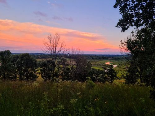 A serene landscape at sunset, featuring rolling hills, a winding river, and colorful skies with scattered clouds.