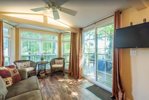 Bright sunroom with a ceiling fan, comfortable seating, and large windows overlooking a deck and greenery outside.