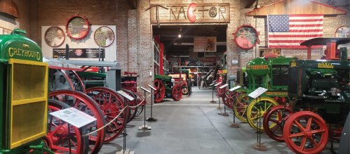 A museum interior showcasing vintage tractors and farming equipment, with brick walls and American flags in the background.