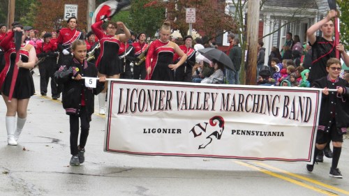 A marching band parade with members in black and red uniforms, holding a banner that reads "Ligonier Valley Marching Band."