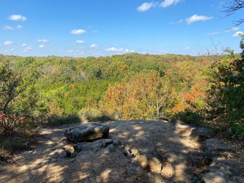 A rocky overlook with a view of colorful autumn trees under a blue sky with scattered clouds.