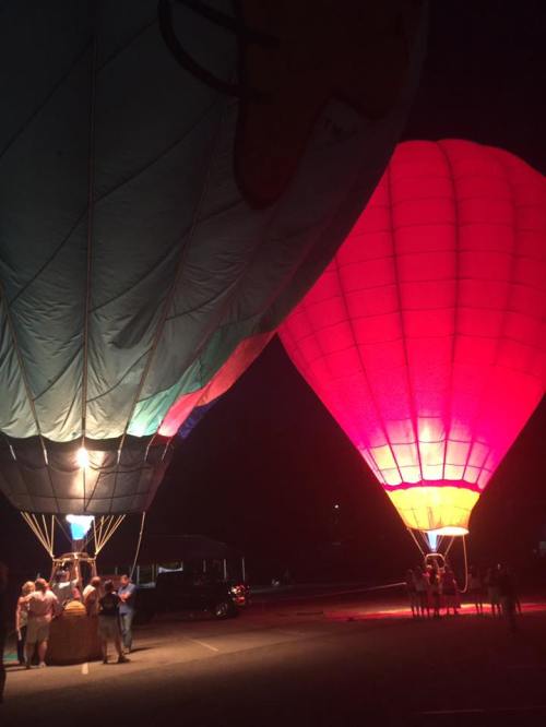 Two brightly lit hot air balloons at night, with people gathered around the bases in a parking area.