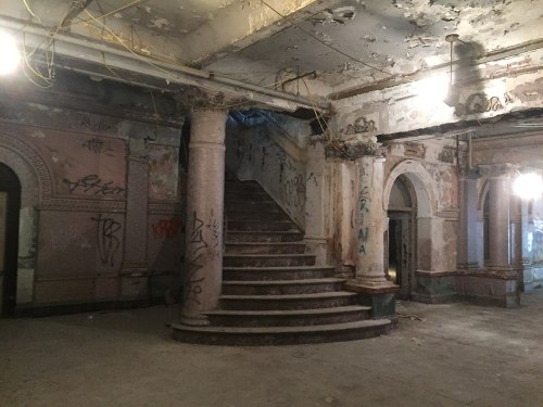 Abandoned interior with peeling walls, graffiti, and a grand staircase leading to an upper level. Dimly lit with exposed wiring.