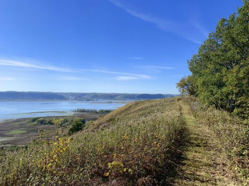 A scenic view of a grassy path along a hillside overlooking a river and distant hills under a clear blue sky.