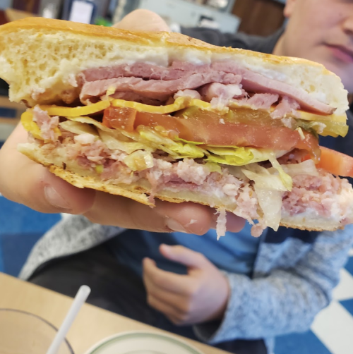 A person holds a large sandwich filled with layers of ham, lettuce, tomato, and other ingredients.