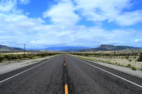 A long, empty road stretches into the distance, flanked by desert landscapes and mountains under a cloudy sky.