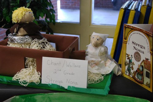 A display featuring two handmade dolls representing characters from "Strega Nona," with a sign for a 2nd-grade project.