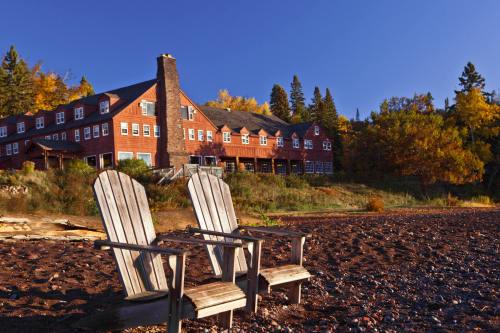 Two wooden chairs on a pebbly beach, with a large lodge and colorful autumn trees in the background under a clear blue sky.