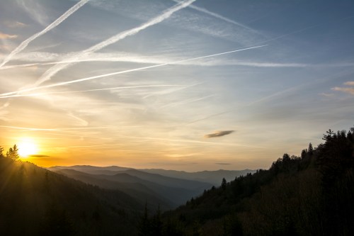 Sunset over a mountainous landscape, with streaks of clouds and a warm glow illuminating the horizon.