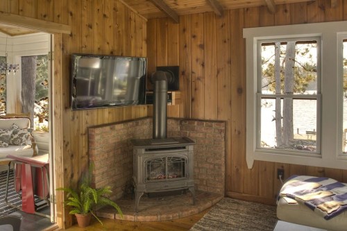 Cozy cabin interior with wooden walls, a brick fireplace, a TV, and large windows overlooking a lake.