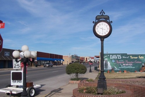 A vintage clock stands on a sidewalk beside a street lined with shops and a speed limit sign in a small town.