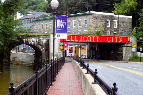 Historic Ellicott City entrance with a stone bridge, red signage, and a scenic view of the river and buildings.