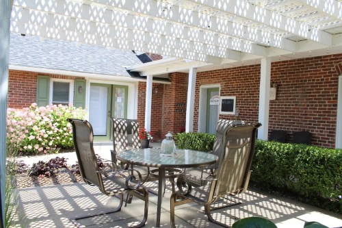 A patio with a table and chairs under a lattice pergola, surrounded by greenery and blooming flowers.