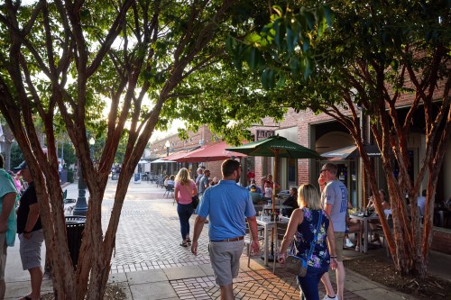 People stroll along a tree-lined street with outdoor dining, enjoying a lively atmosphere during sunset.