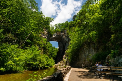 Natural rock arch surrounded by lush greenery and a serene waterway, with people sitting on a bench nearby.