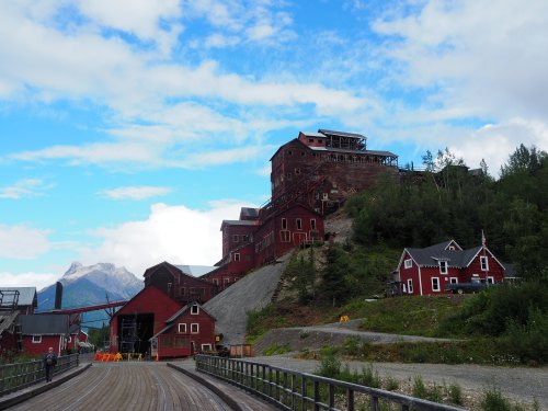 Historic red buildings on a hillside, surrounded by greenery and mountains under a blue sky with scattered clouds.