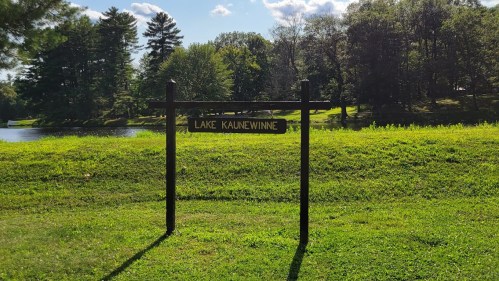 Sign reading "Lake Kaunewinne" in a grassy area near a calm lake, surrounded by trees and blue sky.