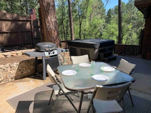 Outdoor patio with a table set for four, a grill, and a hot tub surrounded by trees.