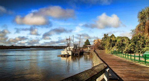 A serene river scene with boats docked, a wooden walkway, and lush greenery under a blue sky with fluffy clouds.
