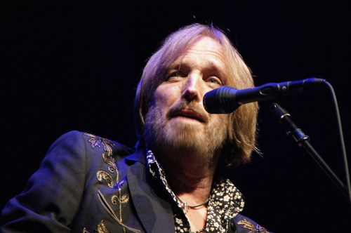 A musician with long hair and a beard sings into a microphone, wearing a stylish jacket with floral designs.