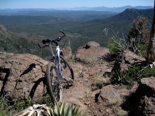 A mountain bike rests on rocky terrain, overlooking a vast forested valley and distant mountains under a clear blue sky.
