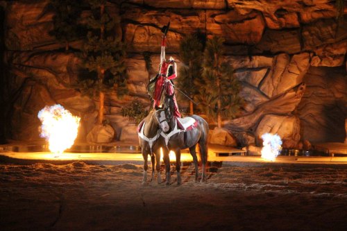 A performer in red attire raises a sword while riding a horse, surrounded by flames and a rocky backdrop.