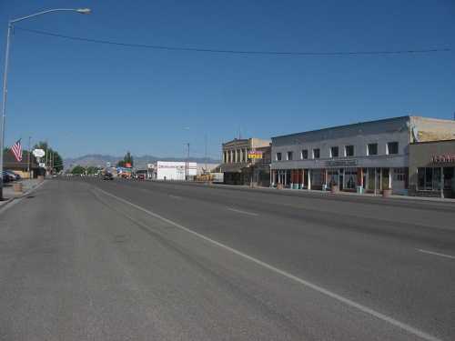 A quiet street scene featuring storefronts and clear blue skies in a small town. Mountains are visible in the distance.