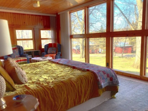 Cozy bedroom with a yellow bedspread, large windows, and a view of trees and cabins outside.