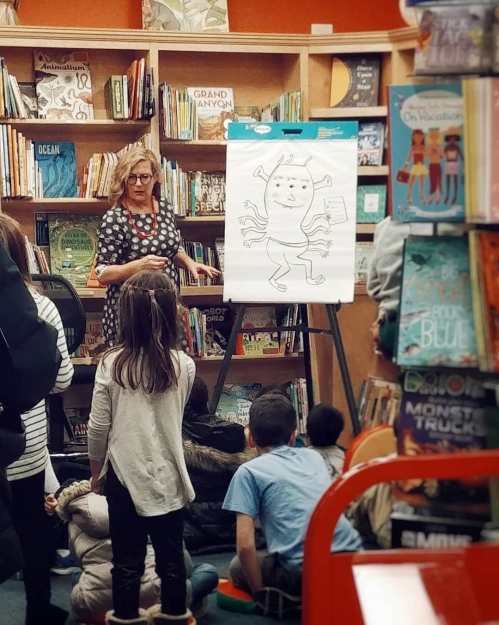 A woman presents a drawing of a monster on an easel to a group of children in a cozy library setting.