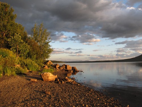 A serene lakeside scene with rocky shore, lush greenery, and dramatic clouds reflecting in the calm water at sunset.