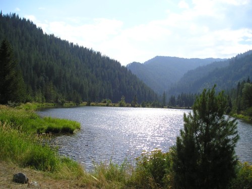 A serene lake surrounded by lush green mountains under a bright sky, reflecting sunlight on the water's surface.