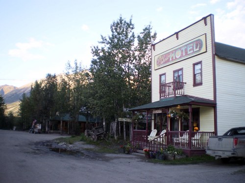 A quaint, yellow hotel with a porch, surrounded by trees and mountains, on a dirt road in a small town.